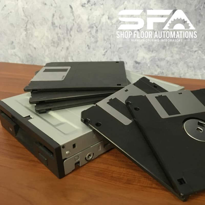 A floppy disk drive on a wooden table with five black floppy disks piled on top and leaning against it.