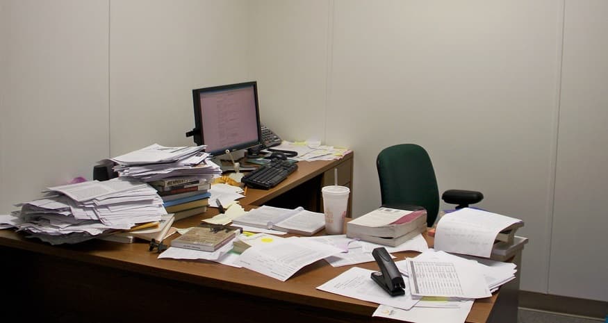 A desk with many piles of paper scattered over it, which can be fixed by going paperless with the use of computers and PDM software.
