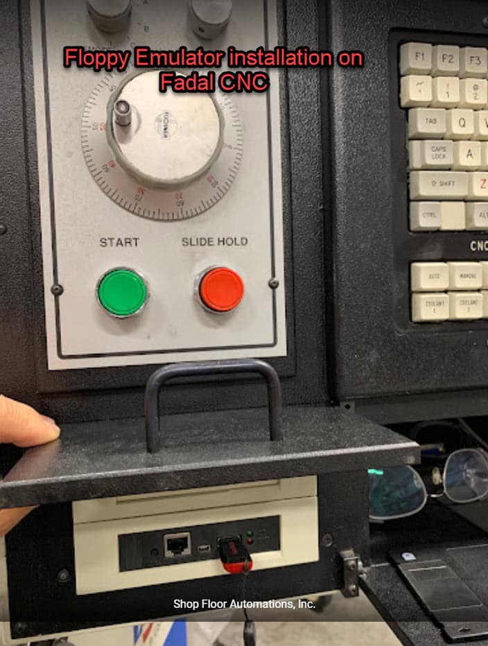 A small door on a Fadal CNC machine is lifted to reveal a Floppy Drive Emulator installed with a USB stick plugged in. The text reads "Floppy Emulator installation on Fadal CNC".