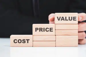 Three stacks of blocks: the shortest stack says "Cost", the middle stack says "Price", and the tallest stack says "Value". A person in the background is holding the "Value" block with two fingers.